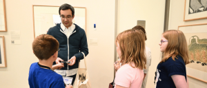 A group of children interact with an adult in an art gallery, discussing an artwork displayed on the wall.
