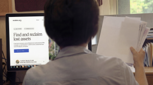 A person, viewed from the back, sits at a computer. The screen shows the "reclaim.org" website with the heading "Find and reclaim lost assets". The person holds a stack of documents.