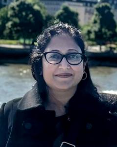 Close-up portrait of Swati in glasses and a dark coat, standing by a riverside promenade with softly blurred buildings and trees in the background.