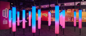 Colorful illuminated columns in a modern exhibit space, featuring vibrant blue and pink lights, with informational displays on the walls.