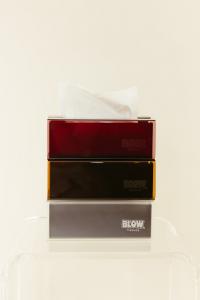 Stacked Blow Tissues refillable acrylic tissue boxes in red, amber, and smoke colors with a tissue pulled through the top box, against a soft neutral backdrop.