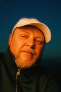 Portrait of musician Damien Cain wearing a light cap against a dark blue background.