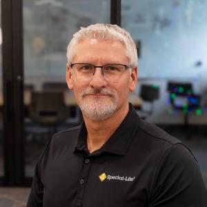 A man in a black polo shirt with glasses and white hair.