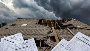 Storm-damaged house roof with missing shingles and exposed rafters under dark clouds, with insurance claim forms in the foreground.