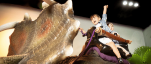 Children joyfully riding a large, dragon-like creature in a playful indoor setting, with bright lights and greenery in the background.