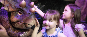 Two children interact with a life-like dinosaur puppet, showing curiosity and excitement in a colorful, adventurous setting.