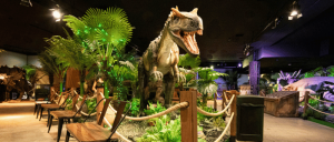 A lifelike dinosaur exhibit featuring a roaring T-Rex amidst lush greenery and wooden seating, creating a prehistoric atmosphere.