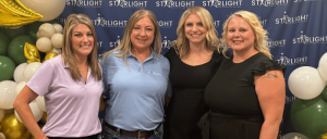 Four women stand together in front of a Starlight-themed backdrop, adorned with balloons in shades of white, green, and gold.