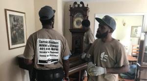 Two Correira Brothers movers in branded uniforms carefully moving an antique grandfather clock in a Rhode Island home