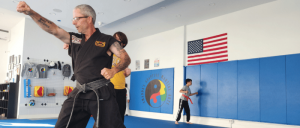 A martial arts class in action, featuring individuals practicing techniques on a blue mat, with American and school flags displayed on the wall.