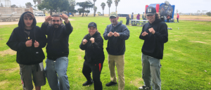 Five individuals in hooded sweatshirts stand in a park, posing with fists raised, surrounded by grass and palm trees.