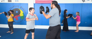 Children practice martial arts techniques in pairs, focused on training with pads in a bright gym setting.