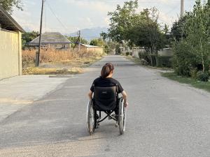A photo of the back of a woman sitting in a wheelchair, She has dark hair in a ponytail, it is daytime and she is in the middle of a road in a rural setting, with mountains in the distance. There is nobody else in the photo, and there are no cars on the r