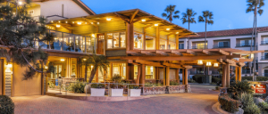 A beautifully lit wooden lodge with large windows and palm trees, showcasing a welcoming entrance at dusk.