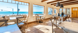 Bright cafe with wooden beams, coastal views, and modern furniture, featuring tables and chairs against large windows overlooking the ocean.