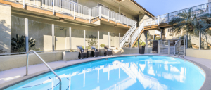 Hotel pool surrounded by lounge chairs, palm trees, and a staircase leading to upper-level rooms under clear blue skies.