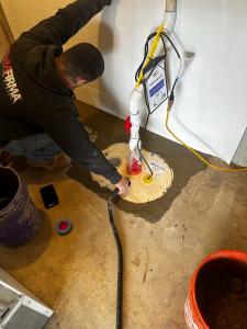 High-angle view of a TerraFirma technician working on a professional sump pump system installed in a basement floor. The system features white PVC discharge pipes and a sealed basin lid surrounded by fresh concrete