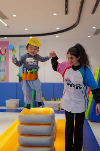 A young child wearing a yellow toy construction hat and tool belt jumps off a soft mat while holding a teacher's hand during a storytelling play session at TuniTuni Tsuen Wan Center, Hong Kong.