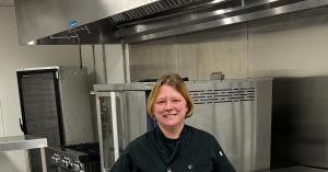 Smiling founder of To the Spoon and Back wearing a chef coat, standing in a commercial kitchen at Frontier Kitchen with stainless steel equipment.