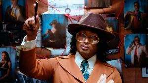 Ashwin Gane wearing a brown suit, tie, and wide-brim hat, holding a pen and standing in front of a corkboard covered with pinned photos and red string connections, portraying a classic investigative setup.