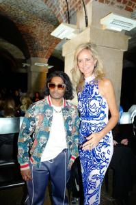 Ashwin Gane in a colorful printed jacket and sunglasses posing with Lady Victoria Hervey in a white and blue patterned dress at a fashion show held in a vaulted, brick-arched venue in London.