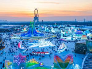 Aerial view of Harbin Ice and Snow World at sunset with large glowing ice sculptures, crowds exploring illuminated structures, and a Ferris wheel in the background.