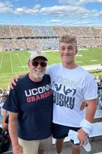 Ryan Corvo, a Confidia Cares scholarship recipient, stands at a University of Connecticut football stadium during a home game, wearing UConn apparel.