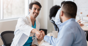 A smiling male healthcare provider in a white coat shakes hands with another professional across a desk during a meeting, symbolizing collaboration, trust, and career partnership in medicine.