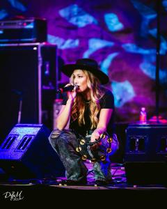 Samantha Rae performing live on stage, crouching with tambourine and microphone under vibrant purple and blue lighting.