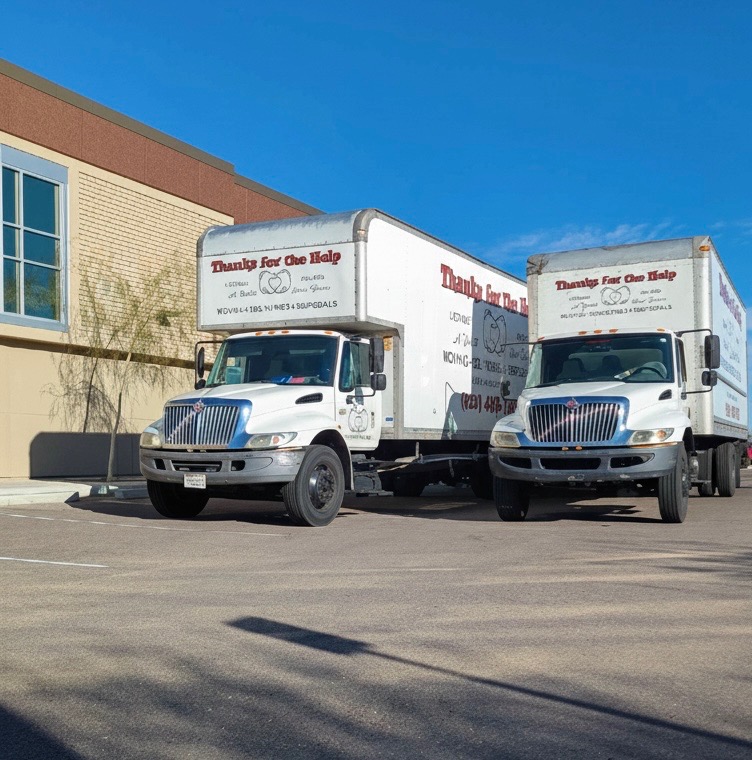 Two white “Thanks for the Help” moving trucks parked side by side in a commercial parking lot near a large building.