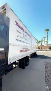 A white “Thanks for the Help” moving truck parked on a residential street with palm trees and homes in the background.