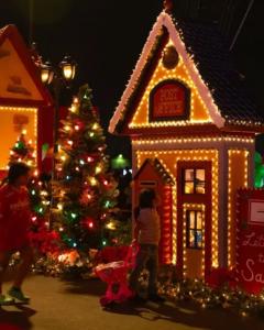 A glowing, miniature holiday post office decorated with bright Christmas lights and a nearby illuminated Christmas tree at night during the Santa Tour event.