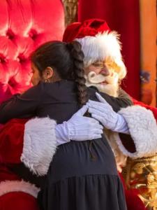 A young girl with a long braid gives Santa Claus a warm hug. Santa is wearing his traditional red suit with white fur and white gloves.