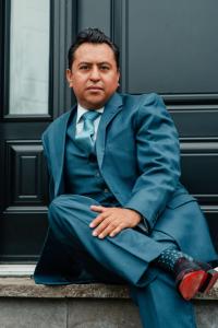 Johnessco Rodriguez wearing a blue suit, seated casually at his residence, reflecting a moment of calm leadership amid the evolving momentum of the international art industry.