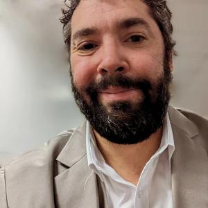 Close-up headshot of a smiling person with dark curly hair and a full beard, wearing a light gray blazer over a white collared shirt, against a softly blurred neutral background.