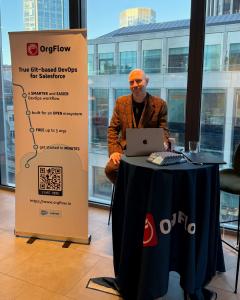 An OrgFlow representative in a brown patterned blazer sits at a conference booth with a laptop. A large vertical banner to his left displays the OrgFlow logo and text reading "True Git-based DevOps for Salesforce" and "FREE up to 3 orgs".