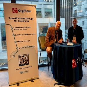 Two men represent OrgFlow at a professional event booth. Behind them, a large banner highlights the platform's benefits, including being "built for an OPEN ecosystem" and offering a "SMARTER and EASIER DevOps workflow".