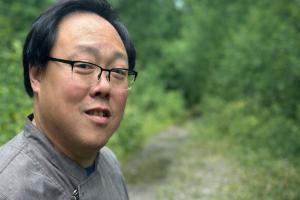 “Portrait of the chef and founder of Wild Onyinz PGH Catering wearing glasses and a gray jacket, standing outdoors on a wooded path with green foliage in the background.”