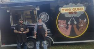 Two owners of Two Gunz BBQ stand in front of their black food truck, smiling and posing outdoors, representing their barbecue food truck business.