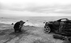 Black and white Image of photographer Mark Maryanovich capturing a burnt car along the PCH next to the ocean.