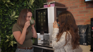 Two employees drinking coffee beside a bean-to-cup coffee machine displaying stories about coffee farmers and impact.