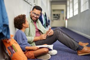 A man and a young student sitting on the floor of a school hallway, smiling and talking together, with an open book in the student's lap.