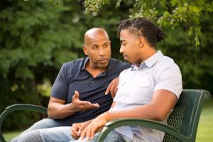 An older man talking with a younger man on a park bench, his hand on the younger man's shoulder in a supportive gesture.