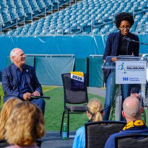 A professional speaker, Nile Livingston, Founder and CEO of Creative Repute, stands at a podium with a microphone inside a stadium, with rows of empty seats visible in the background, representing large-scale venues used for public events and announcement