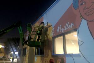 Artist on a scaffold outlining a projected image with paint to start a mural