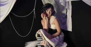 Alisha Liston sitting on set in a purple skirt, white blouse and black and white stockings holding a pomegranate in her hand