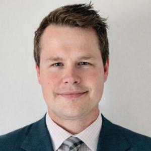 Professional headshot of Hazen Baron, general counsel of Coinme, wearing a dark suit jacket, light patterned dress shirt and tie, facing the camera with a neutral, approachable expression against a clean, light background.