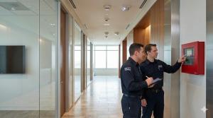 Two uniformed men work with a fire alarm panel in a modern, bright hallway. One points at the screen while the other takes notes on a clipboard.
