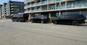 Three black luxury vehicles, including a Mercedes Sprinter and two Cadillac Escalades, parked in front of a modern building.