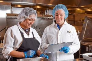 Two women wearing hair nets and gloves inspect a meal in a commercial kitchen.